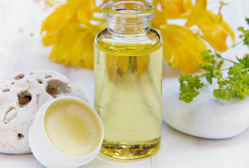 Eine Glasflasche mit gelber Flüssigkeit steht neben einem kleinen offenen Gefäß mit Balsam auf einer Oberfläche. In der Nähe befinden sich ein strukturierter Stein, ein glatter weißer Stein, ein Zweig mit grünen Blättern und verschwommene gelbe Blumen im Hintergrund.