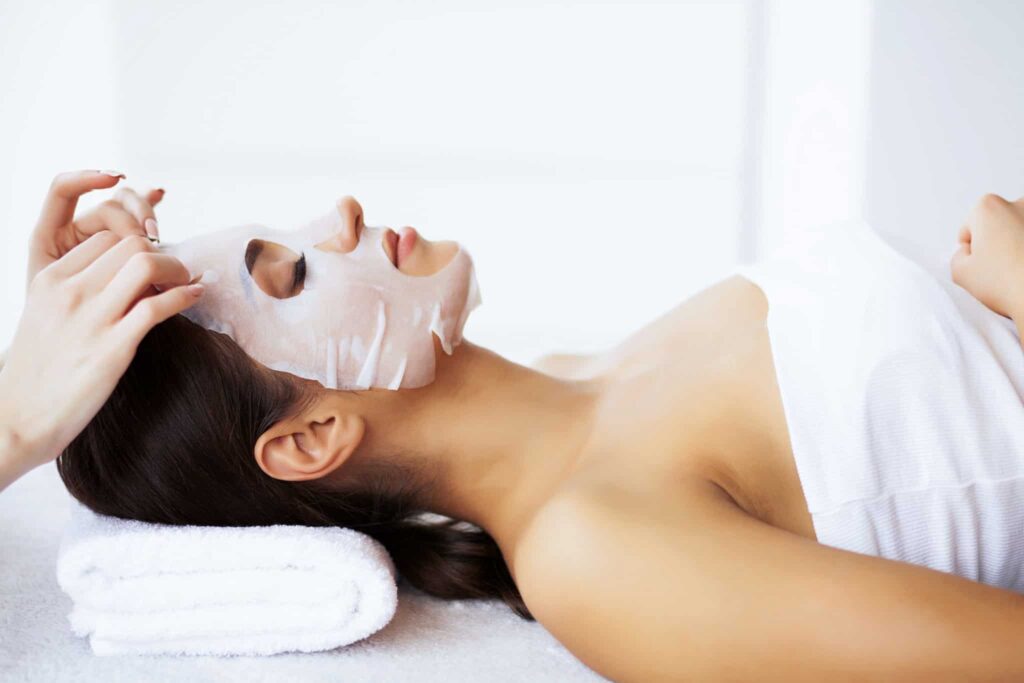 Eine Person liegt mit geschlossenen Augen da und genießt eine kosmetische Gesichtsbehandlung. Sie trägt eine weiße Gesichtsmaske. Eine andere Person rückt die Maske sanft auf ihr Gesicht zu. Sie ruht auf einem gefalteten weißen Handtuch und ihre Schultern sind mit einem weichen weißen Handtuch oder Kleidungsstück bedeckt.