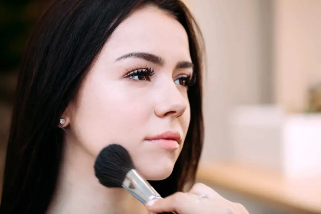 Eine Person mit langen dunklen Haaren lässt sich mit einem Pinsel kunstvoll Make-up auf das Gesicht auftragen. Der Hintergrund ist sanft unscharf und hebt so den Fokus auf das Gesicht der Person.