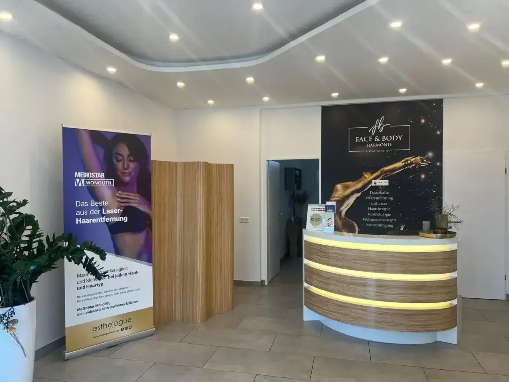 Empfangsbereich mit einem geschwungenen weißen Schreibtisch, Deckenleuchten und Werbebannern für Schönheitsbehandlungen, Kosmetologie und Wellness-Massagen.