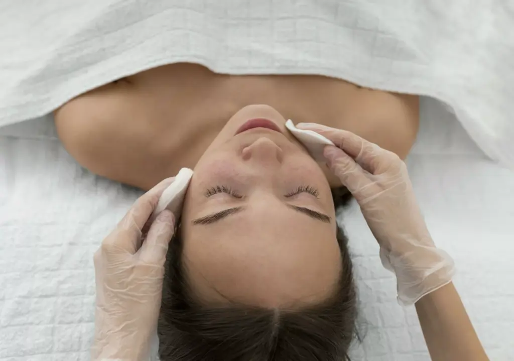 Eine Person erhält eine Gesichtsbehandlung in einem Spa oder einer Hautpflegeklinik in Ulm. Behandschuhte Hände legen sanft Wattepads auf ihr Gesicht, während sie auf einem mit einem weißen Handtuch bedeckten Tisch liegt und sich einer wohltuenden Hauttherapie hingibt.