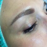 Nahaufnahme des geschlossenen Auges einer Person. Man sieht gepflegte Augenbrauen und lange Wimpern. Die Haut erscheint glatt und auf der linken Seite des Bildes ist teilweise eine grüne OP-Haube zu sehen.